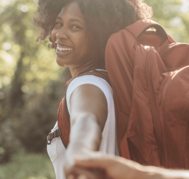 A black woman with a large rucksack holding a man’s hand and laughing, in nature, sun shining from behind