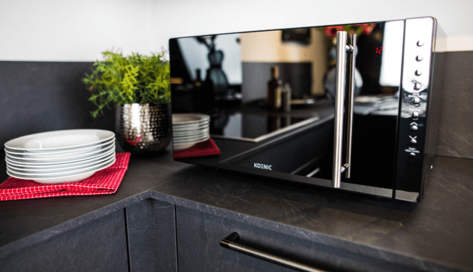 Mikrowelle der Marke KOENIC auf einer Küchentheke, dunkle Marmortextur, daneben ein Stapel weißer Teller, Mikrowelle reflektiert Küche