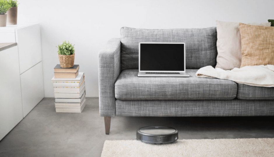 Stofzuigrobot van het merk KOENIC zuigt stof in een moderne woonkamer, laptop ligt op een bank, panorama