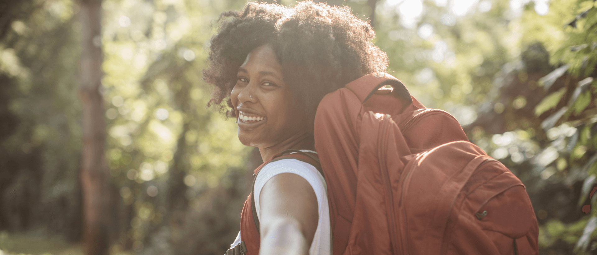 Mulher negra com uma grande mochila a sorrir, segurando a mão de um homem, na natureza, sol a brilhar por trás