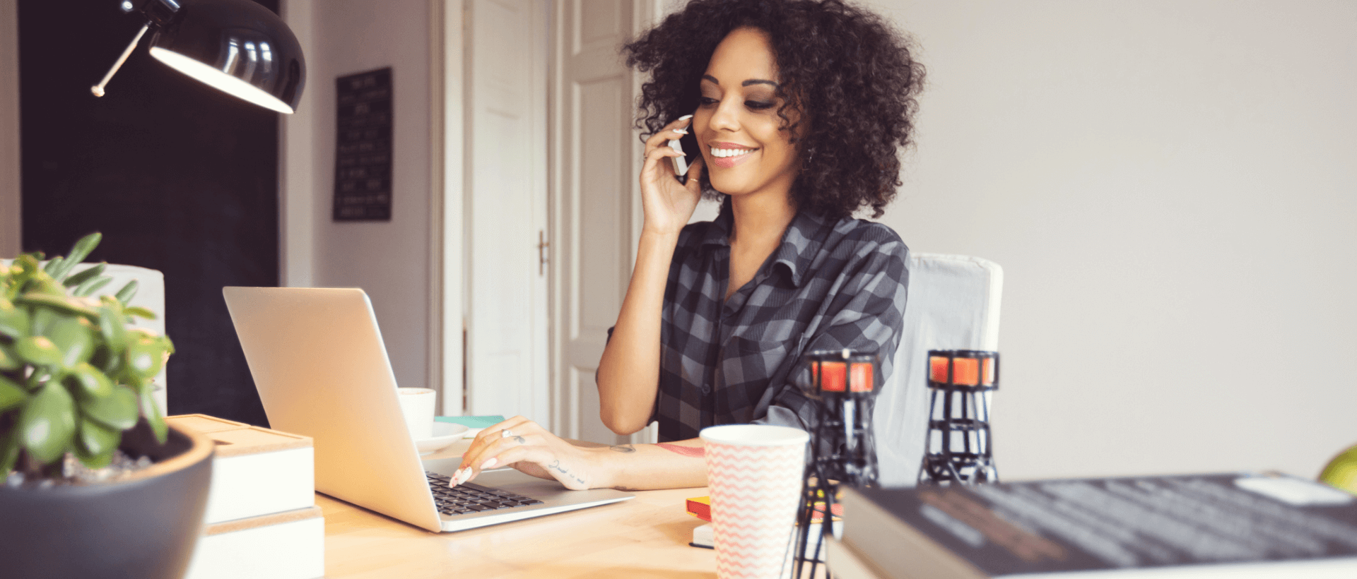 Uma mulher jovem faz uma chamada telefónica com o seu smartphone no seu local de trabalho, com a outra mão escreve no seu portátil, num ambiente de escritório